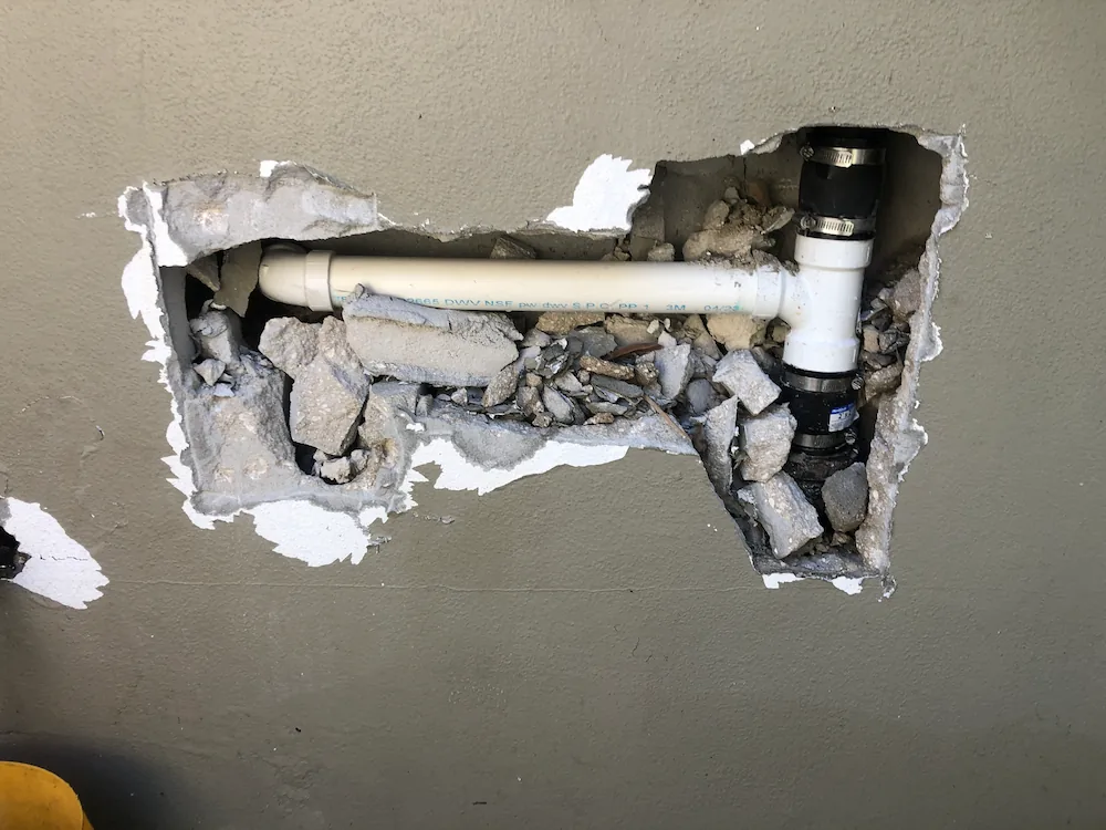 Hole in a gray wall exposing white PVC plumbing pipes surrounded by broken concrete and debris.