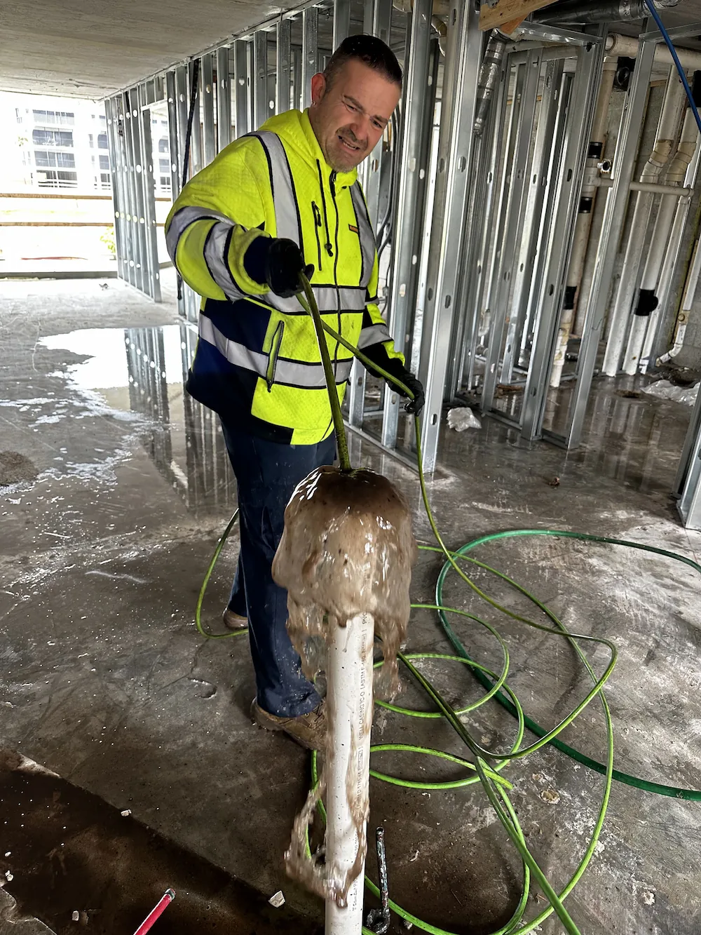 Construction worker in a high-visibility jacket clearing a clogged white pipe with rising muddy water in an unfinished building interior.