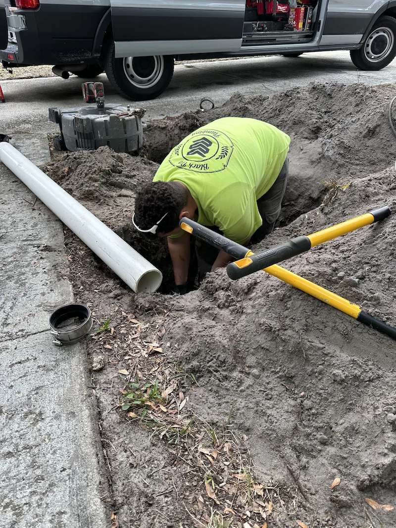 Worker in a neon yellow shirt kneeling in a dirt hole beside a large white pipe and yellow-handled tools, with a van in the background.