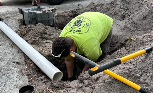 Worker in a neon green shirt digging in a trench next to a white pipe and a yellow-handled digging tool lying on the ground.