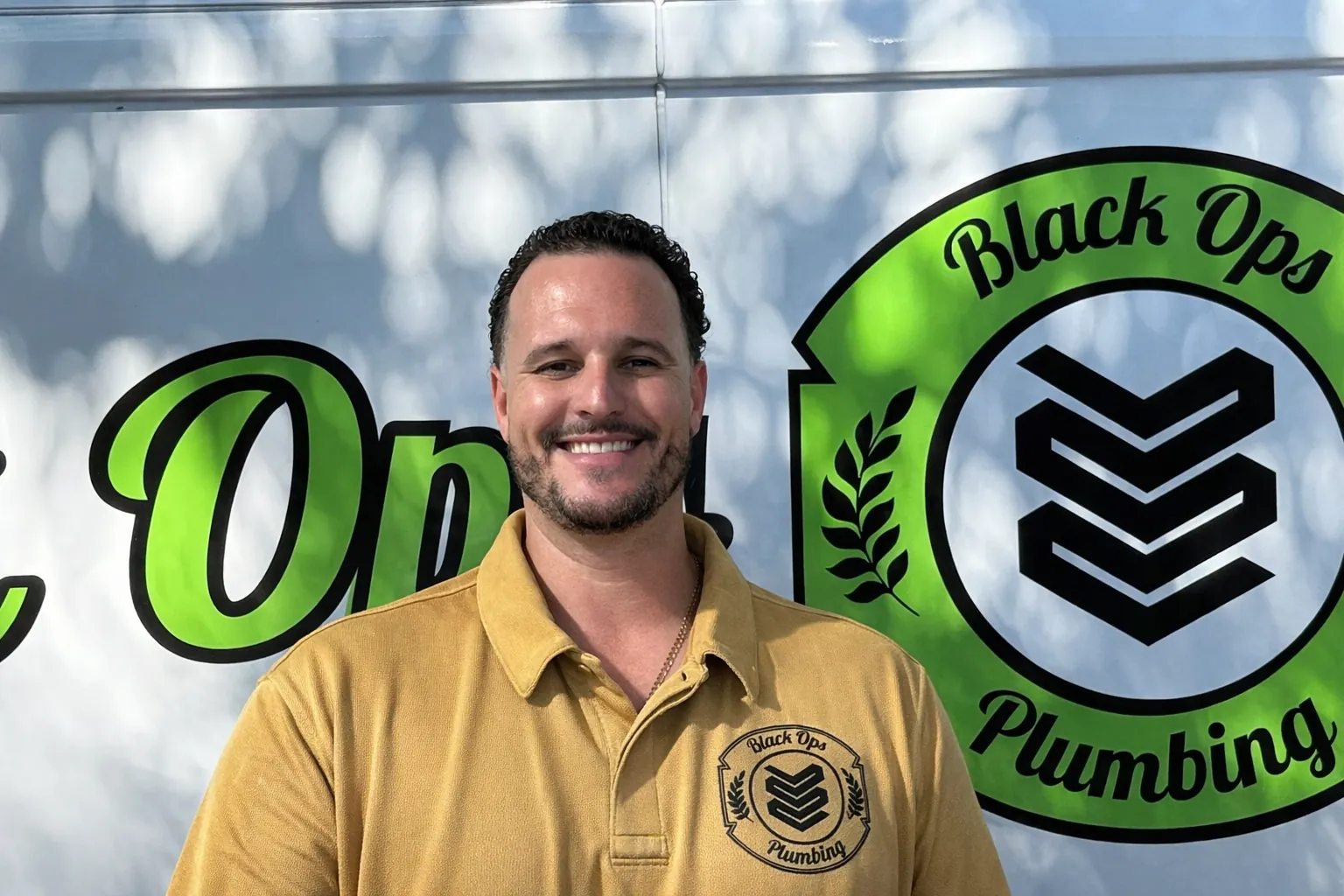 Smiling man wearing a yellow Black Ops Plumbing shirt standing in front of a vehicle with Black Ops Plumbing logo.