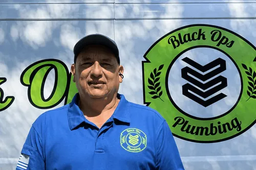 Man wearing a blue polo shirt and cap standing in front of a Black Ops Plumbing logo on a truck.