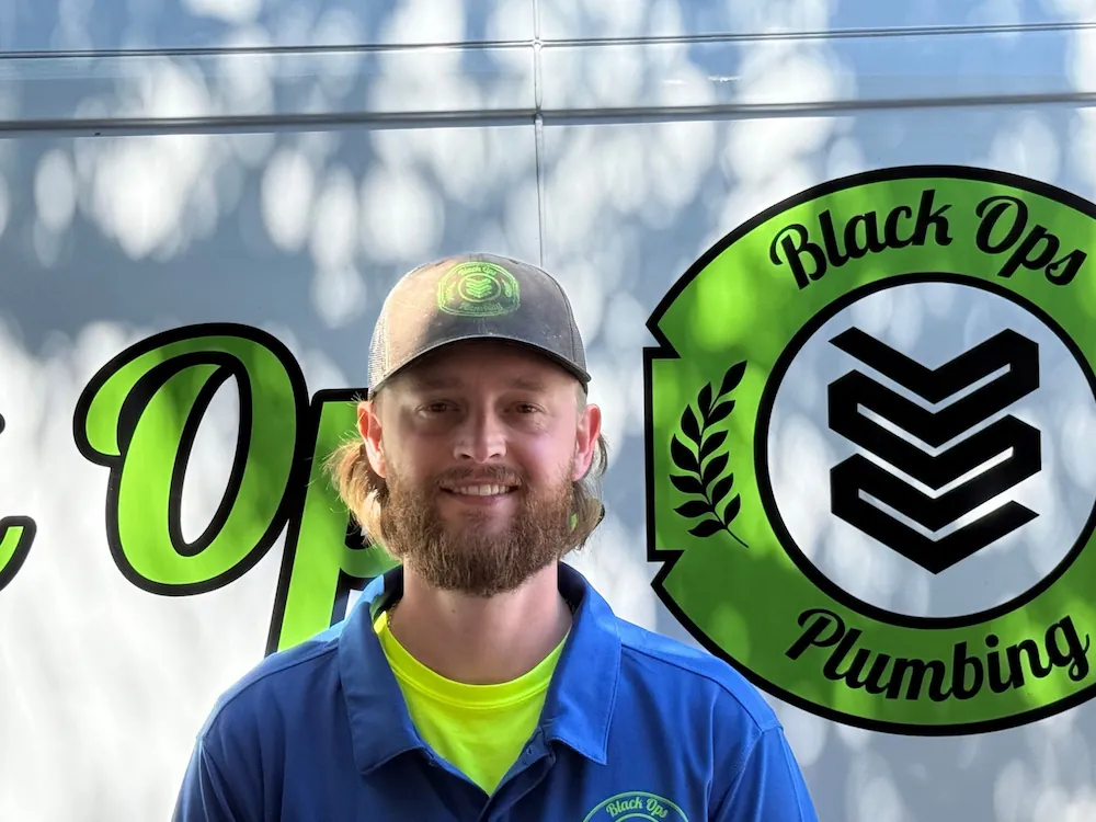 Smiling man with beard wearing a Black Ops Plumbing uniform and cap standing in front of a Black Ops Plumbing logo.