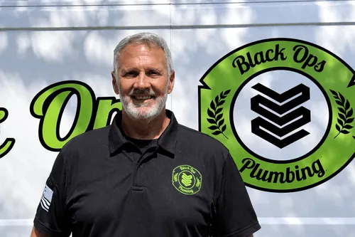 Smiling man with gray hair and beard wearing a black Black Ops Plumbing polo shirt standing in front of a branded service van.