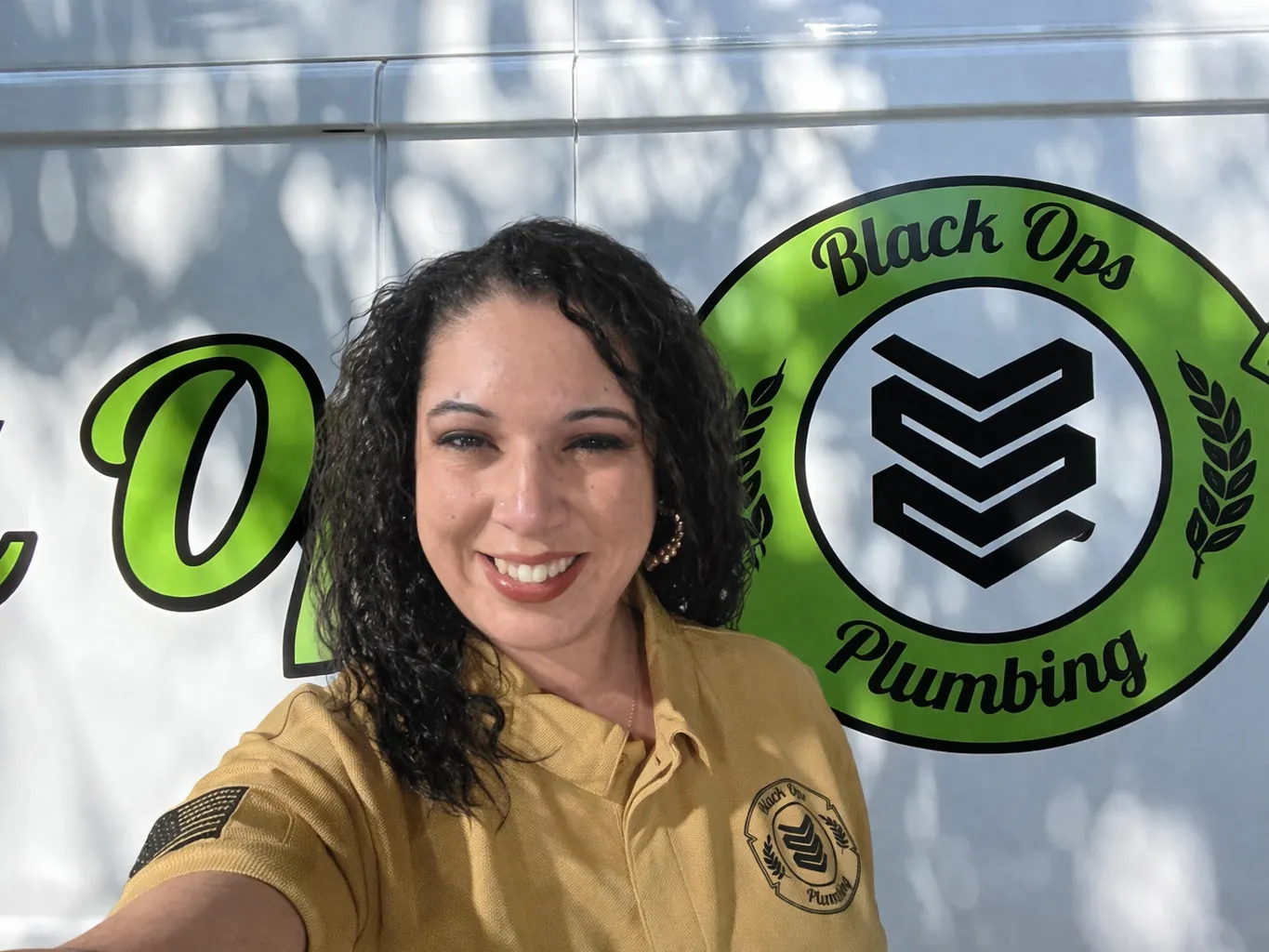 Woman with curly hair wearing a tan Black Ops Plumbing uniform shirt, smiling and standing in front of a vehicle with the company logo.