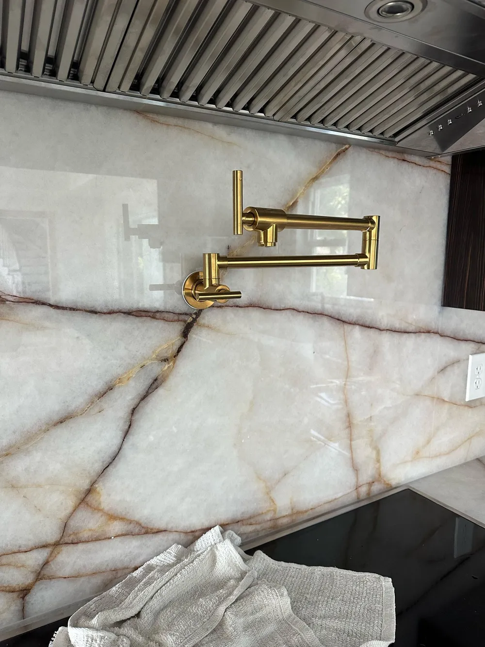 Brushed gold pot filler faucet mounted on a white marble kitchen backsplash with a vent hood above and white cloths on a black stovetop below.