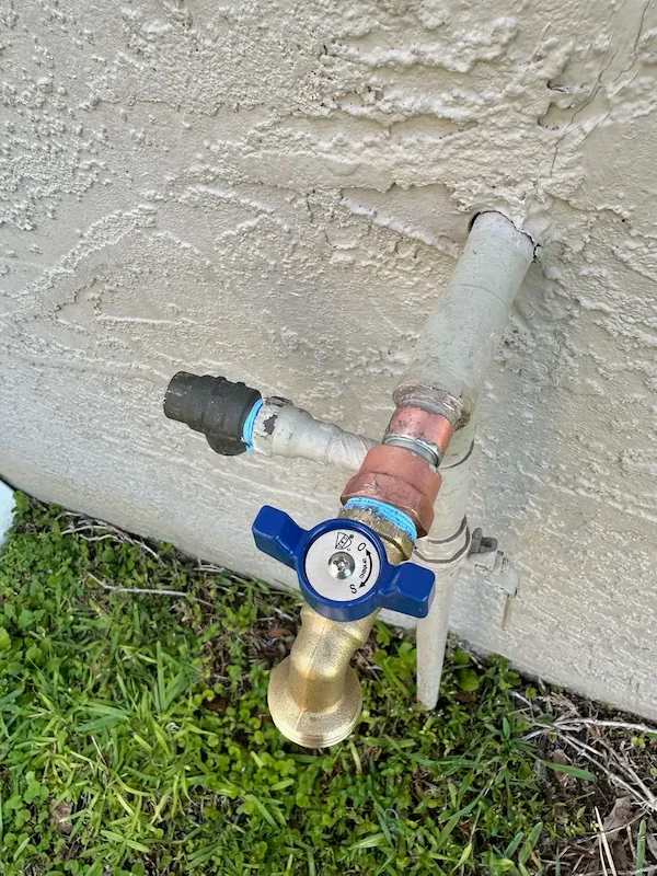 Outdoor water spigot with a blue handle attached to a textured beige wall above green grass.
