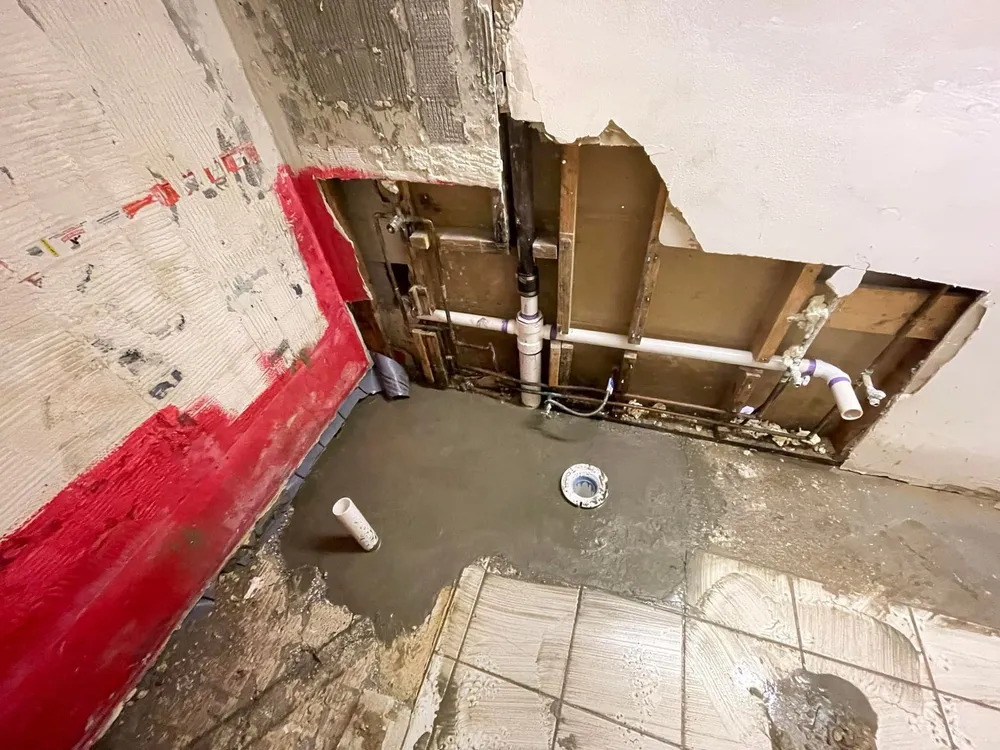 Bathroom under renovation with exposed pipes in the wall, wet cement on the floor around drain pipes, and partially removed tiles and wall covering.