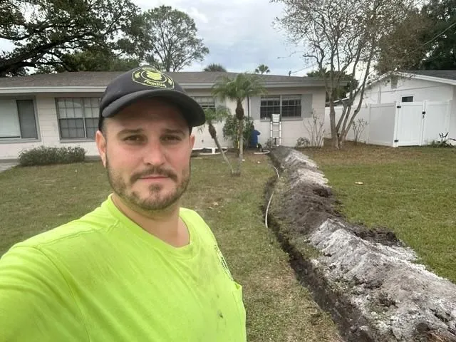 Man in a bright green shirt and black cap taking a selfie in front of a trench dug in a lawn beside a house.