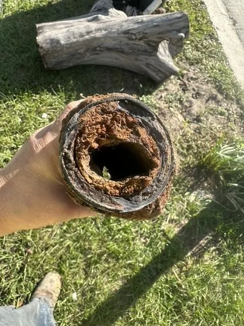Hand holding a heavily rusted and corroded metal pipe with a hollow center, outdoors on grass near a tree log.