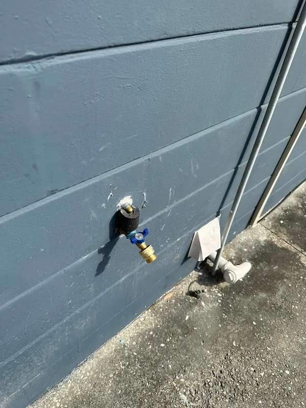 Outdoor water faucet with blue handle attached to a gray concrete wall next to a white pipe.
