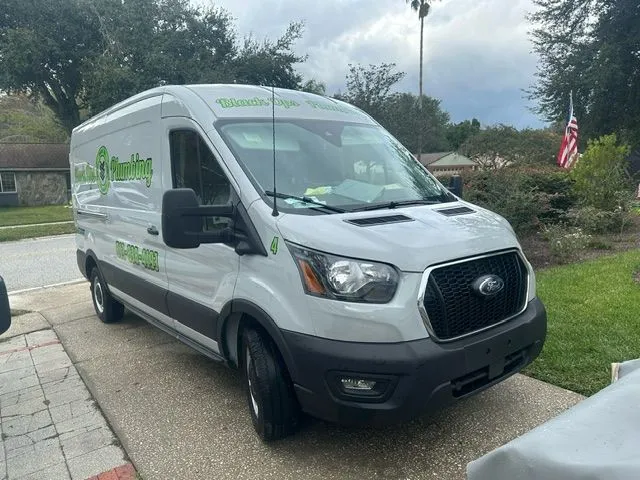White Ford transit van parked on a residential driveway with green 'Plumbing' logo and contact number on the side.
