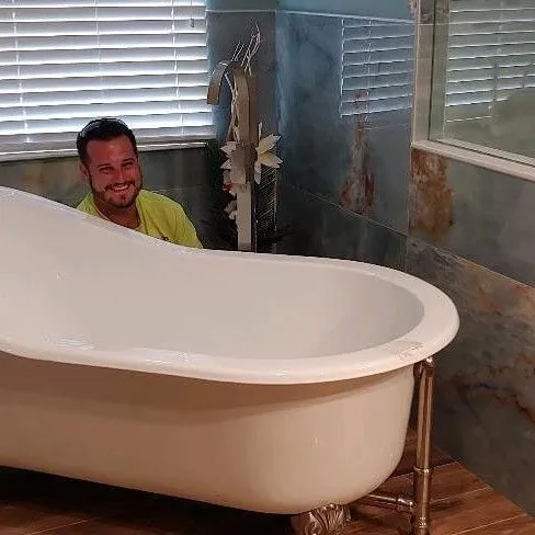 Man smiling while partially hidden behind a white freestanding bathtub in a bathroom with tiled walls and a window with blinds.