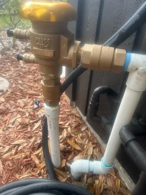 Brass valve assembly connected to white PVC pipes and black hoses near a dark wall with mulch ground covering.