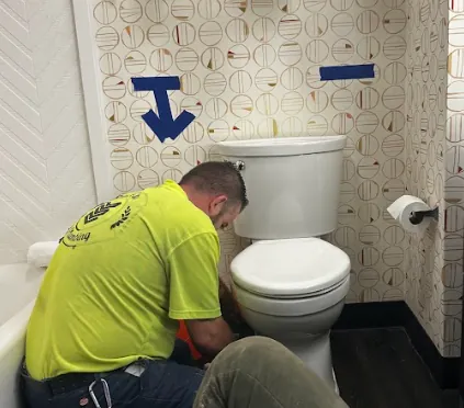Man in yellow shirt working on a white toilet in a bathroom with patterned walls and a blue arrow pointing down.