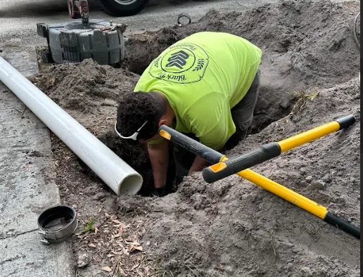 Worker in a bright green shirt digging a trench beside a sidewalk with large yellow-handled tools and a white pipe nearby.