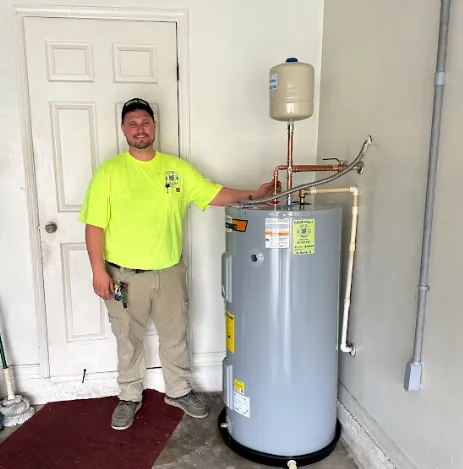 Man in a yellow shirt and beige pants standing next to a gray water heater in a corner of a room.