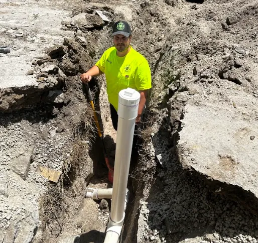 Worker wearing a neon yellow shirt and cap standing in a trench next to white PVC pipes for plumbing or drainage.