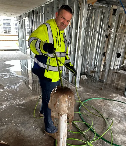 Construction worker wearing a high-visibility jacket using a hose to spray water on a pipe inside an unfinished building.
