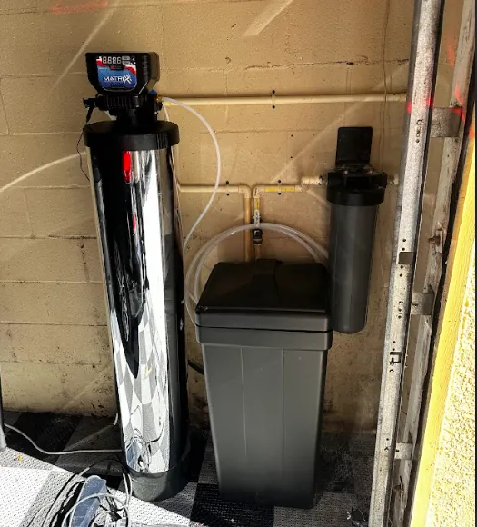 Home water softener system consisting of a tall chrome tank, a gray brine tank, and a black water filter mounted on a beige wall.