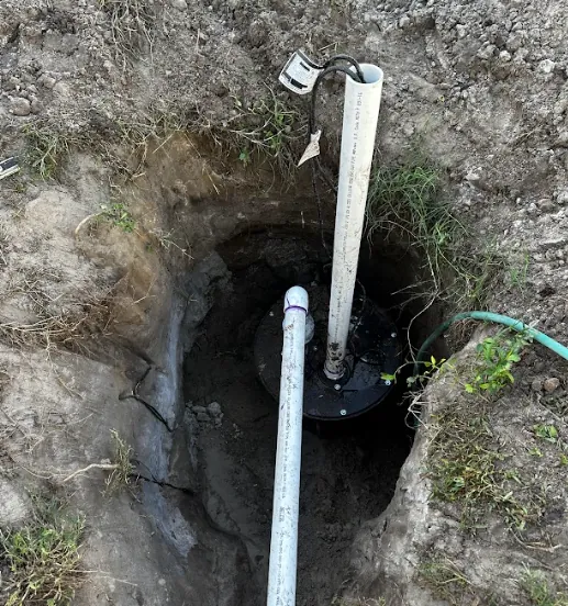 A sump pump installed in a deep hole in the ground with two white PVC pipes connected to it, surrounded by dirt and some grass.