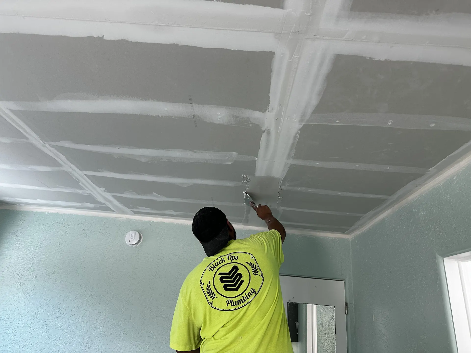 Person in a neon yellow Black Ops Plumbing shirt smoothing drywall on a ceiling in a room with light blue walls.