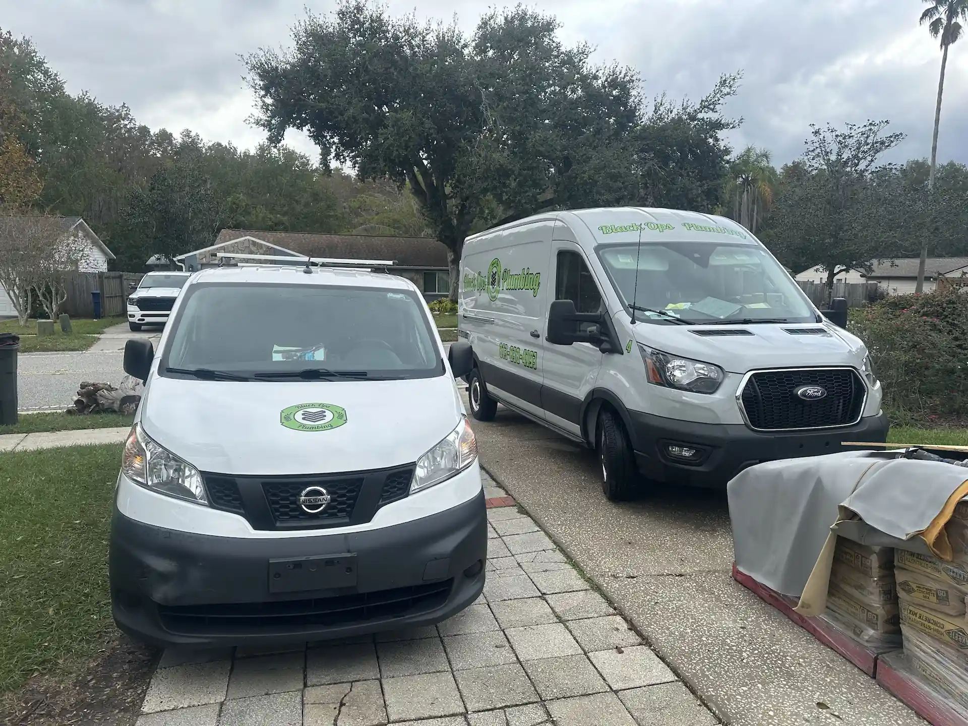 Two white work vans parked outside a house, one with Black Ops Plumbing logo and contact number on the side.