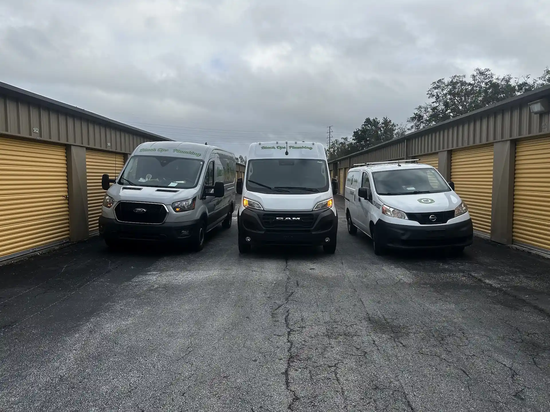 Three white service vans parked between yellow storage unit doors under a cloudy sky.