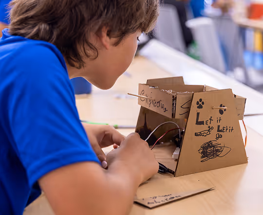 Boy working on a cardboard electronics project labeled 'Let it go' on a classroom table.