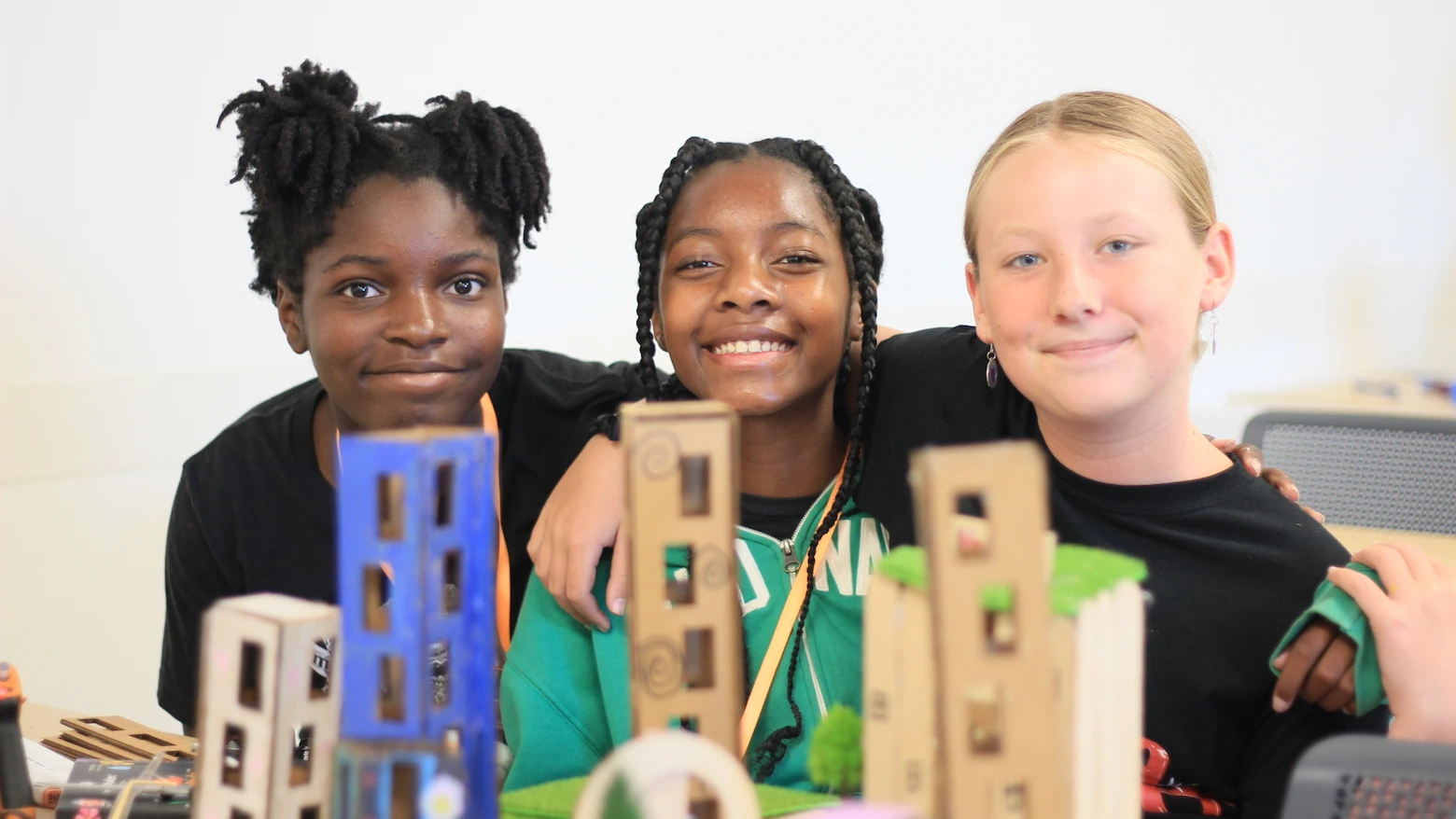 Three smiling children with arms around each other behind a table displaying small model buildings.