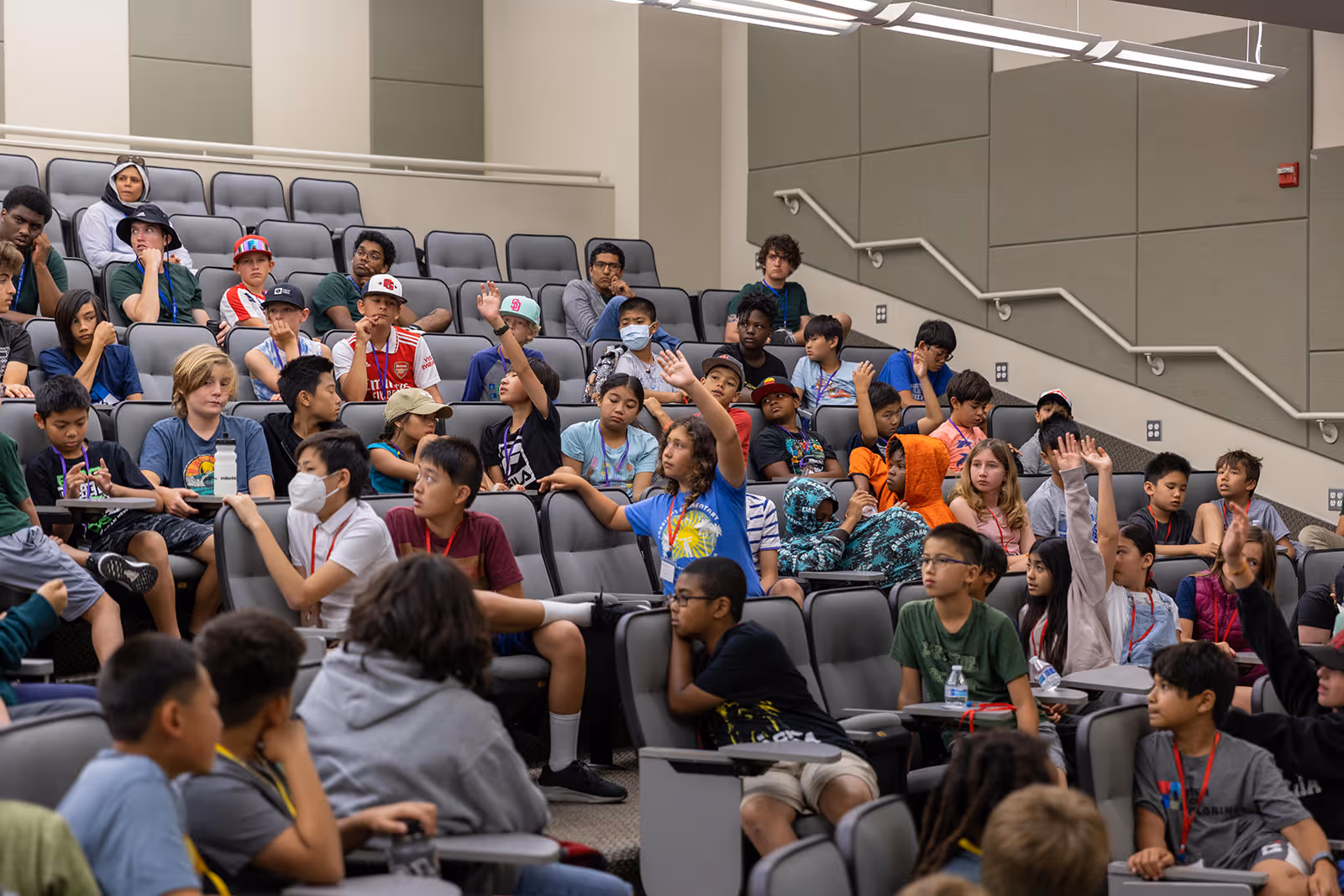 A diverse group of children seated in a tiered lecture hall, some raising their hands.