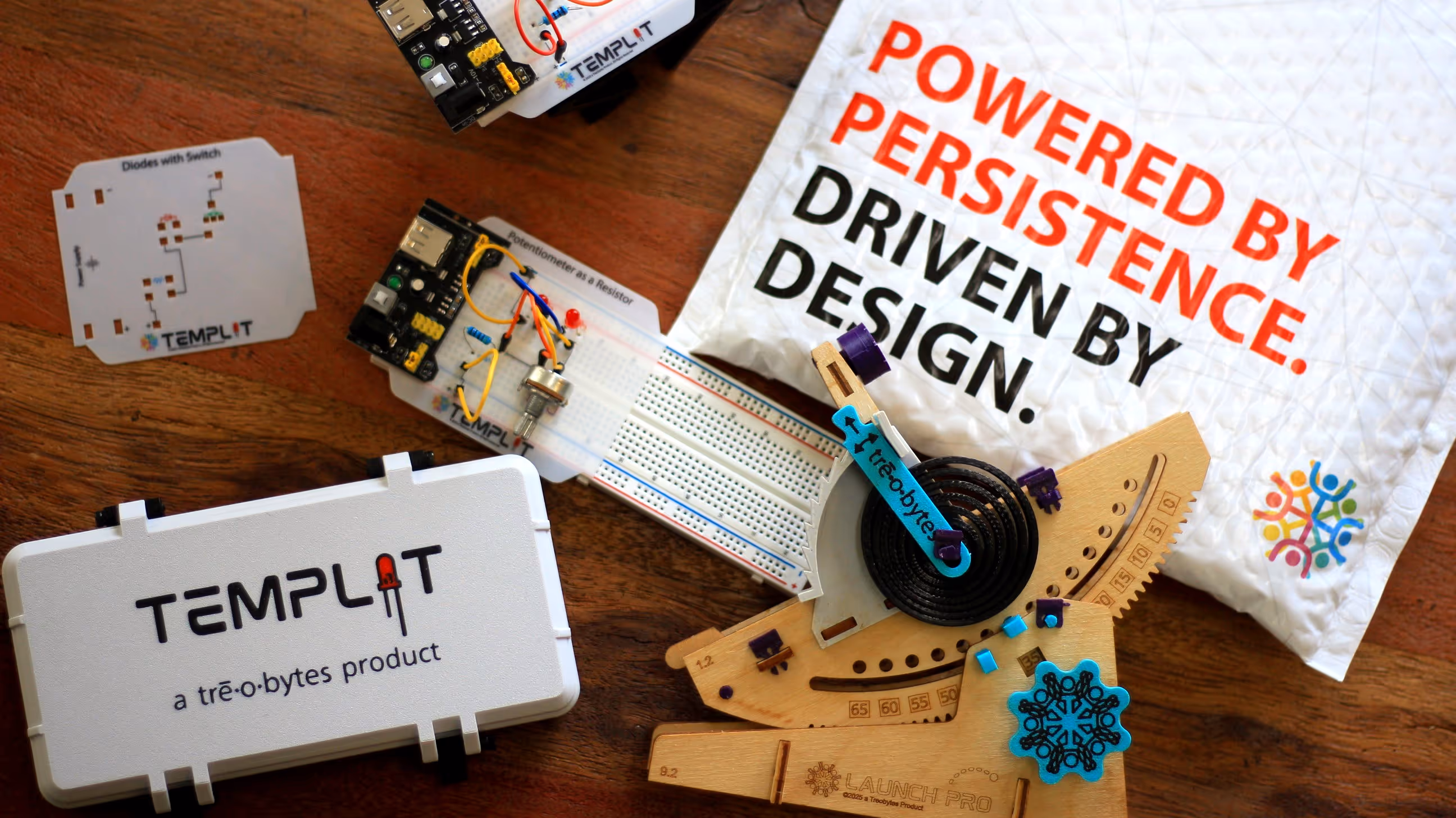 Wooden Launch Pro mechanical device next to Templit electronics kit and white bag with slogan 'POWERED BY PERSISTENCE. DRIVEN BY DESIGN.'