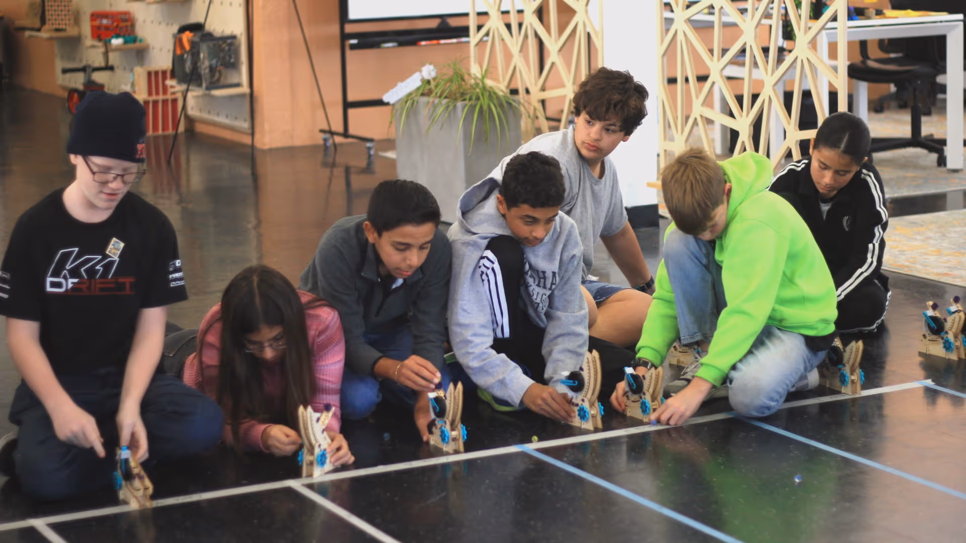 Six children seated on the floor assembling small wooden mechanical devices lined up along a taped grid on a shiny black floor.