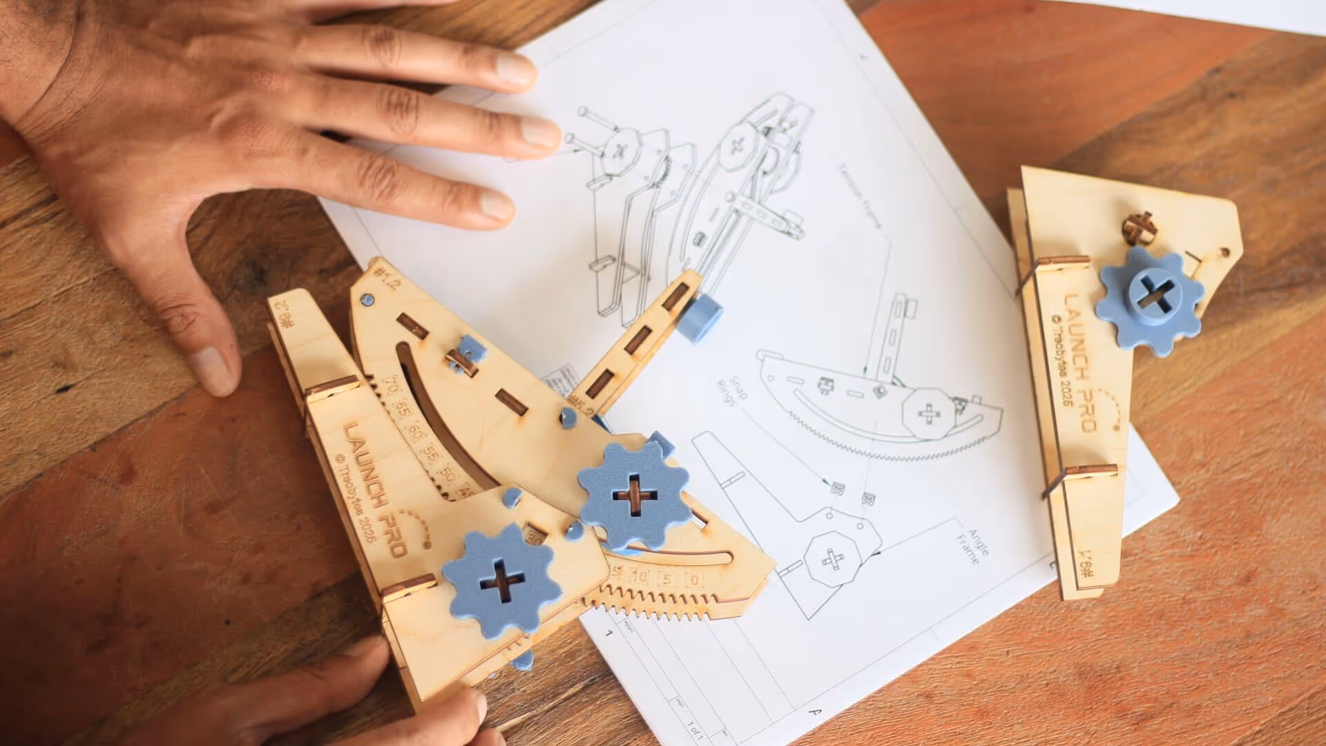 Hands assembling a wooden mechanical model labeled 'Launch Pro' on a wooden table with assembly instructions beneath.