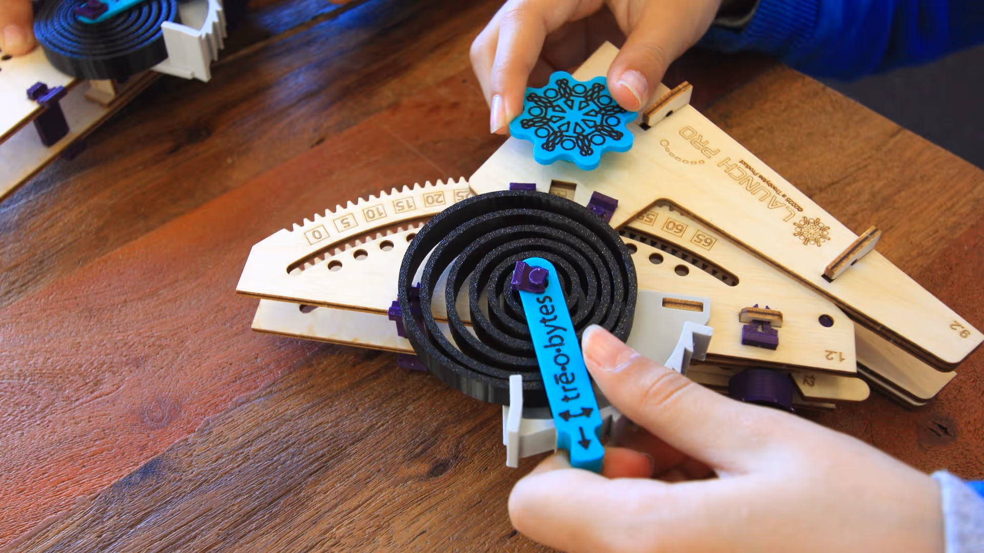 Hands adjusting gears on a wooden Launch Pro mechanical device on a wooden table.
