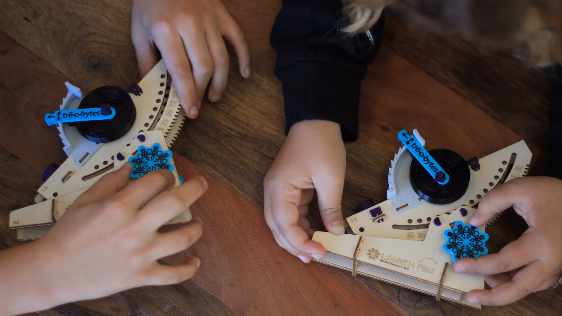 Two children assembling or adjusting wooden mechanical model pieces on a wooden table.