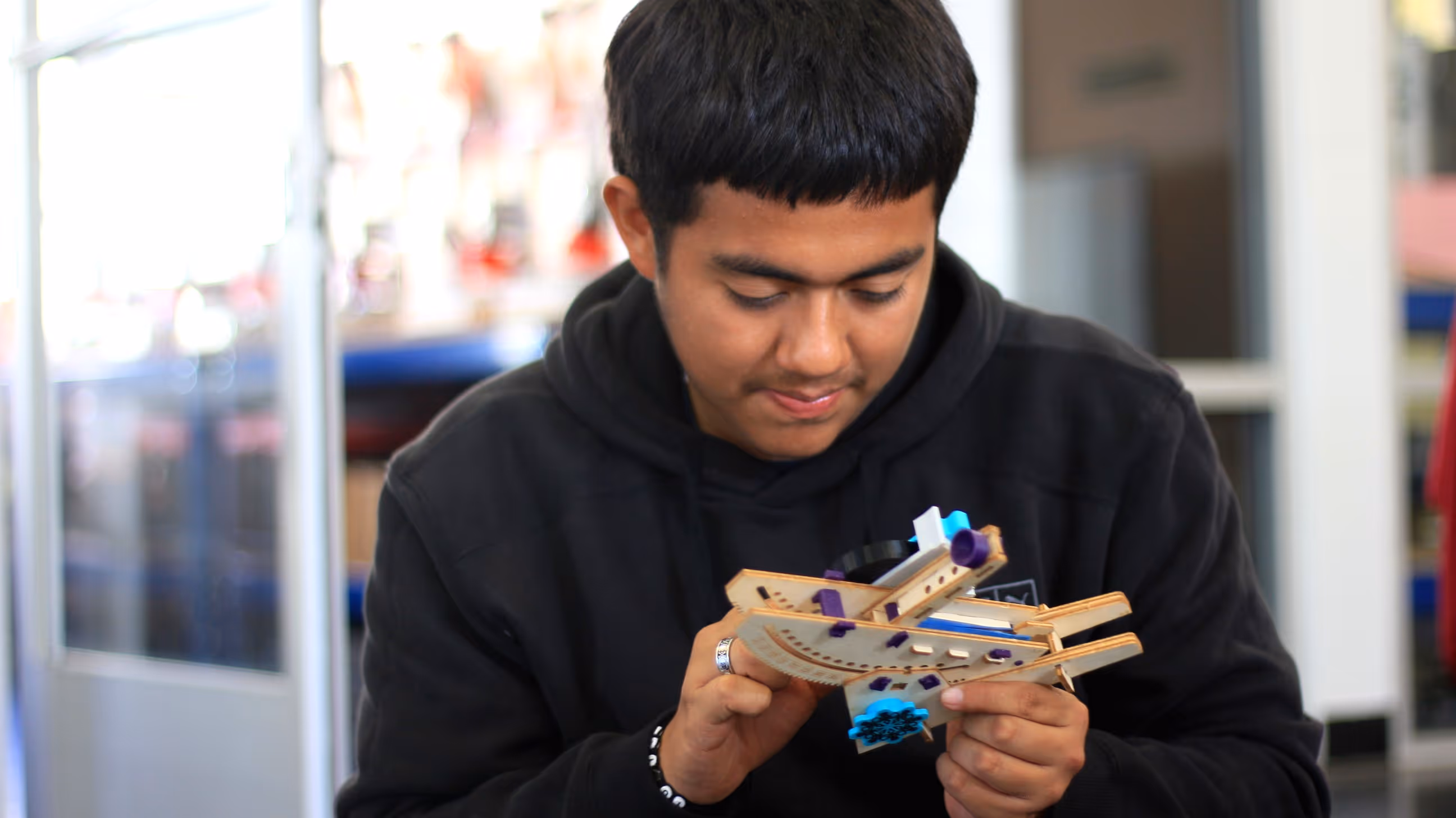Young man examining a small wooden mechanical model with purple and blue components indoors.