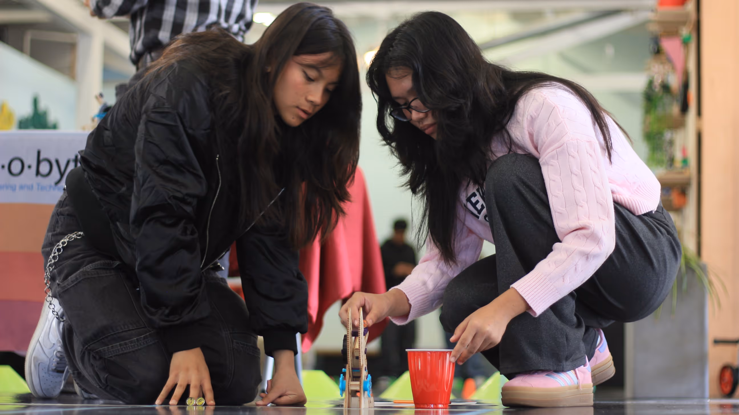 Two young women kneeling on the floor, examining a small wooden mechanical device next to a red cup.