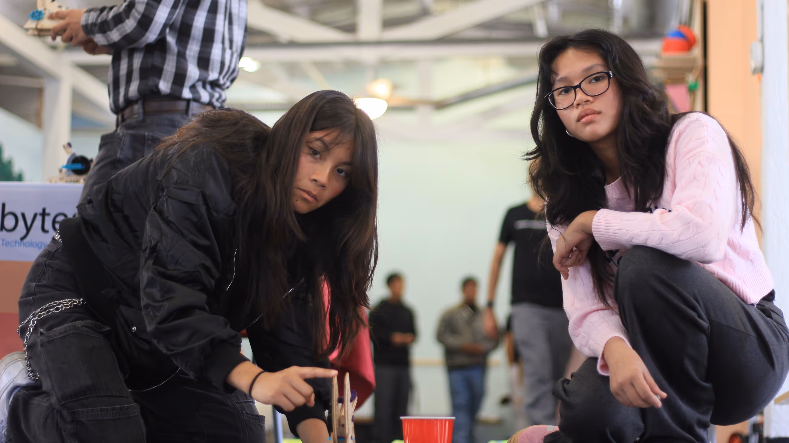 Two young women crouching indoors, one pointing at a wooden mechanical object while the other watches attentively.