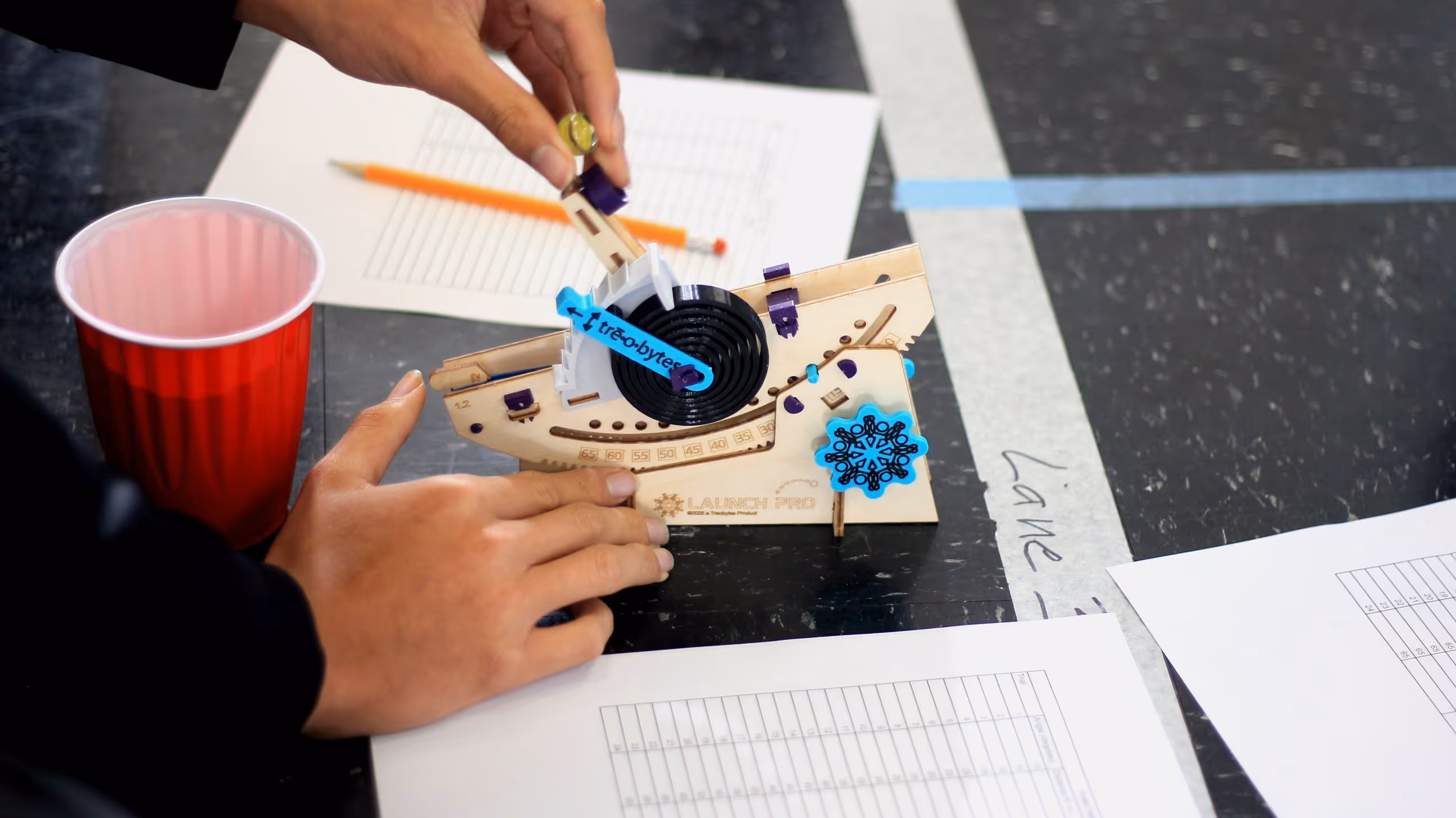 Hands assembling a wooden Launch Pro mechanical device with blue accents on a tabletop with papers, pencil, and a red plastic cup nearby.