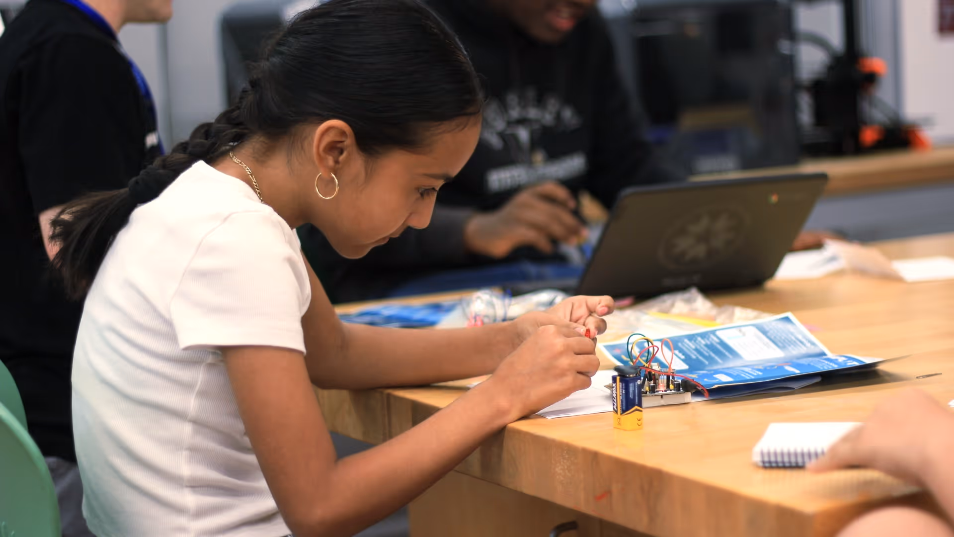 Young girl working on an electronics project with a battery and wires at a wooden table, with a laptop and other students in the background.