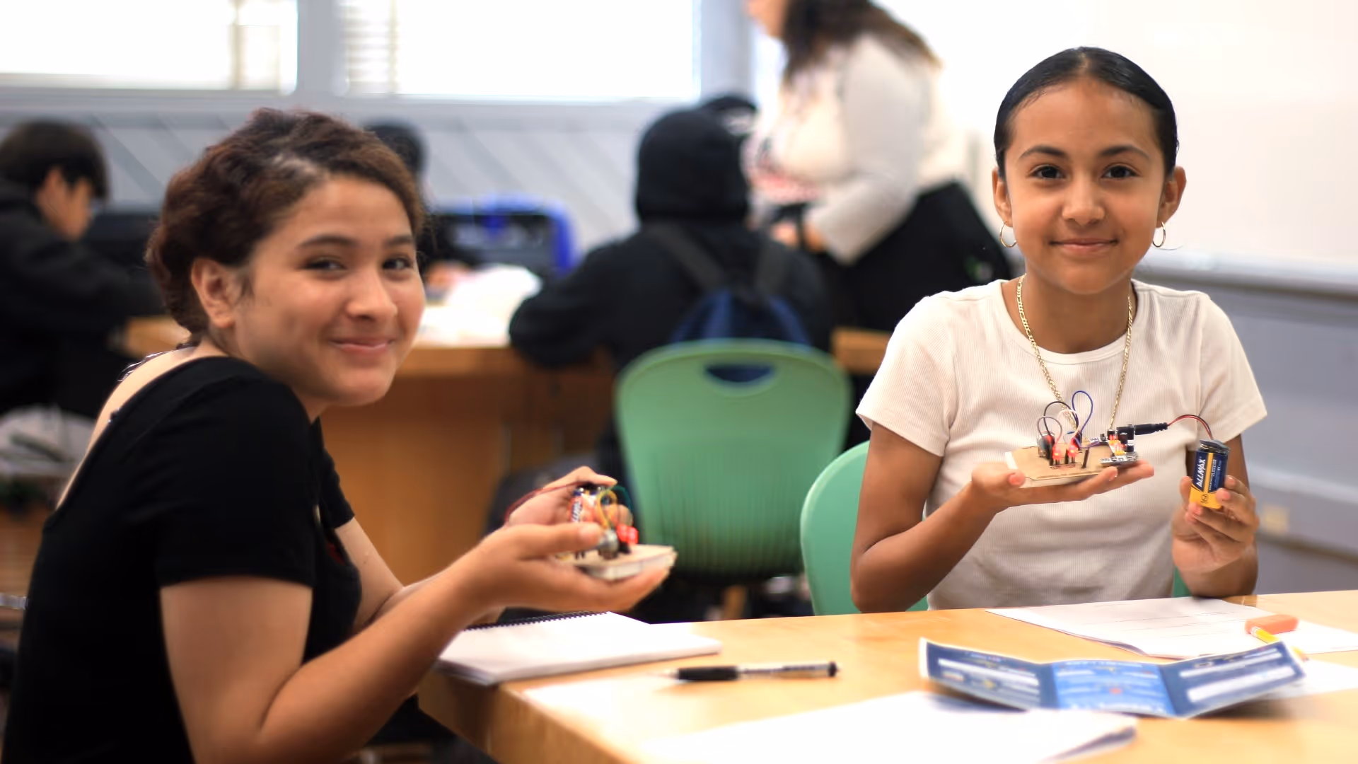 Two girls sitting at a table holding small electronic circuit projects with wires and a battery.