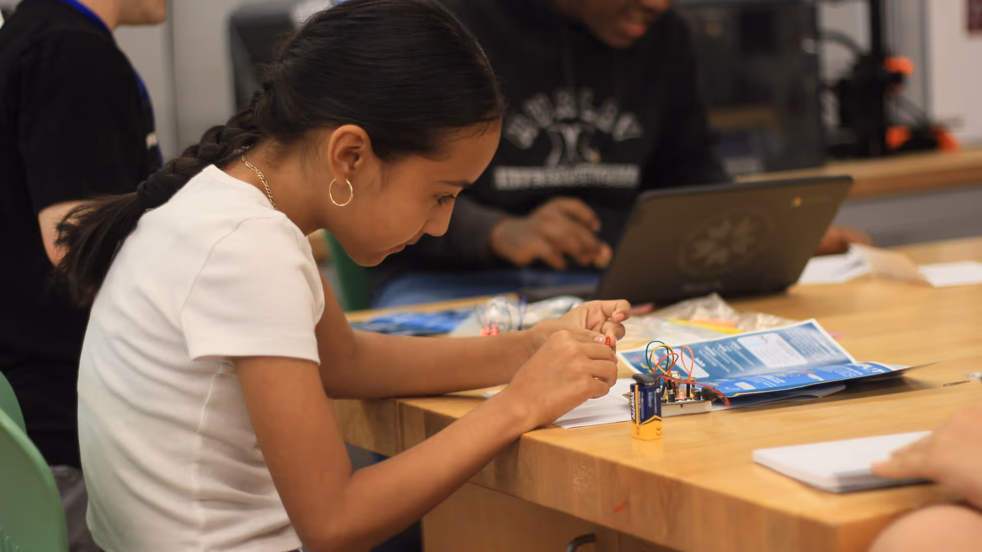 Young girl with braided hair working on an electronics project at a wooden table with a battery and wires.