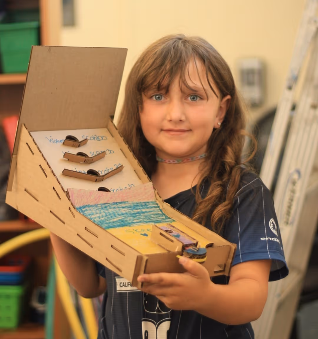 Young girl holding a homemade cardboard skee-ball game with colored sections and ball ramps.