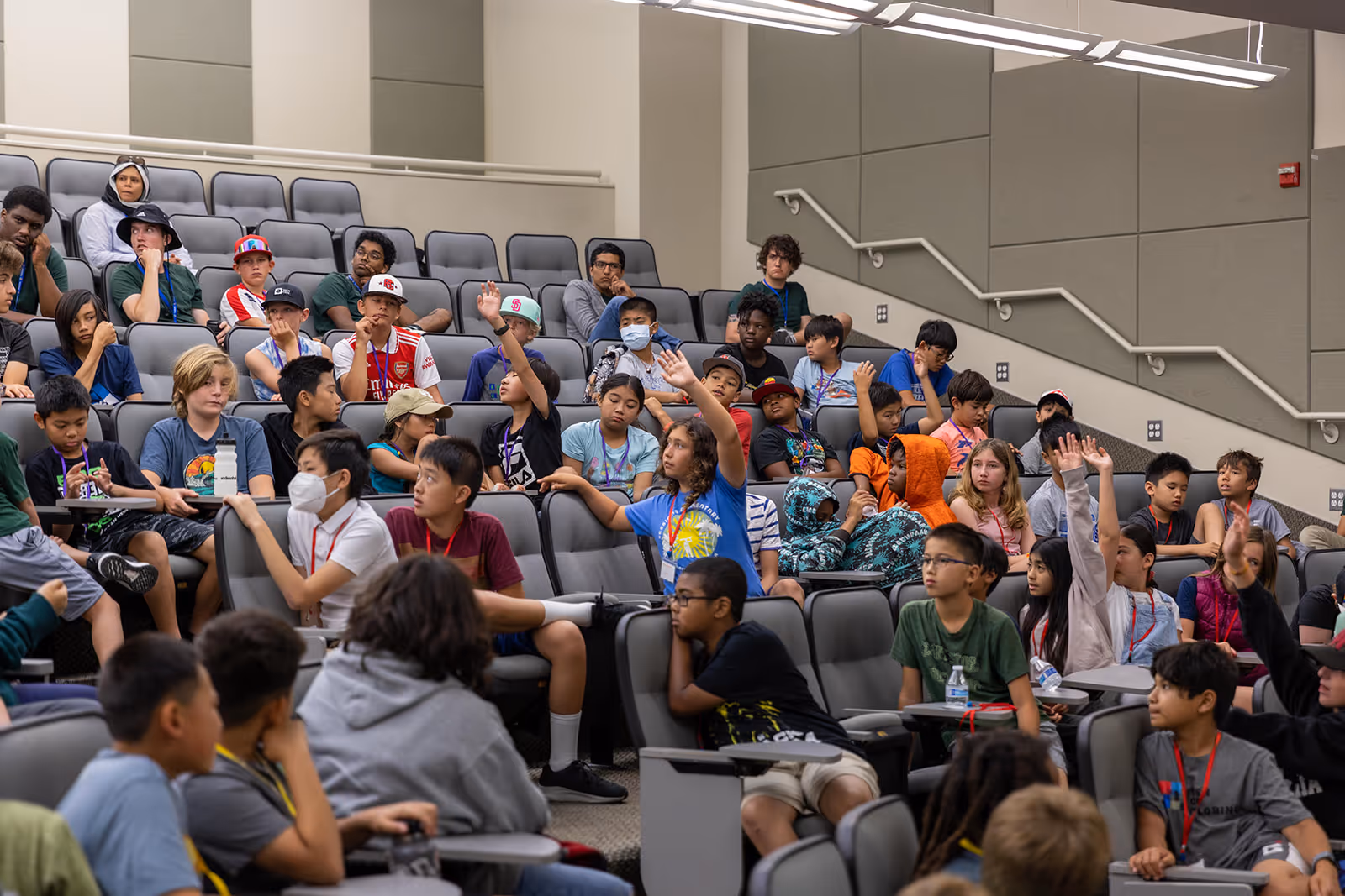 A group of children seated in an auditorium, some raising their hands, engaged in a discussion or lecture.