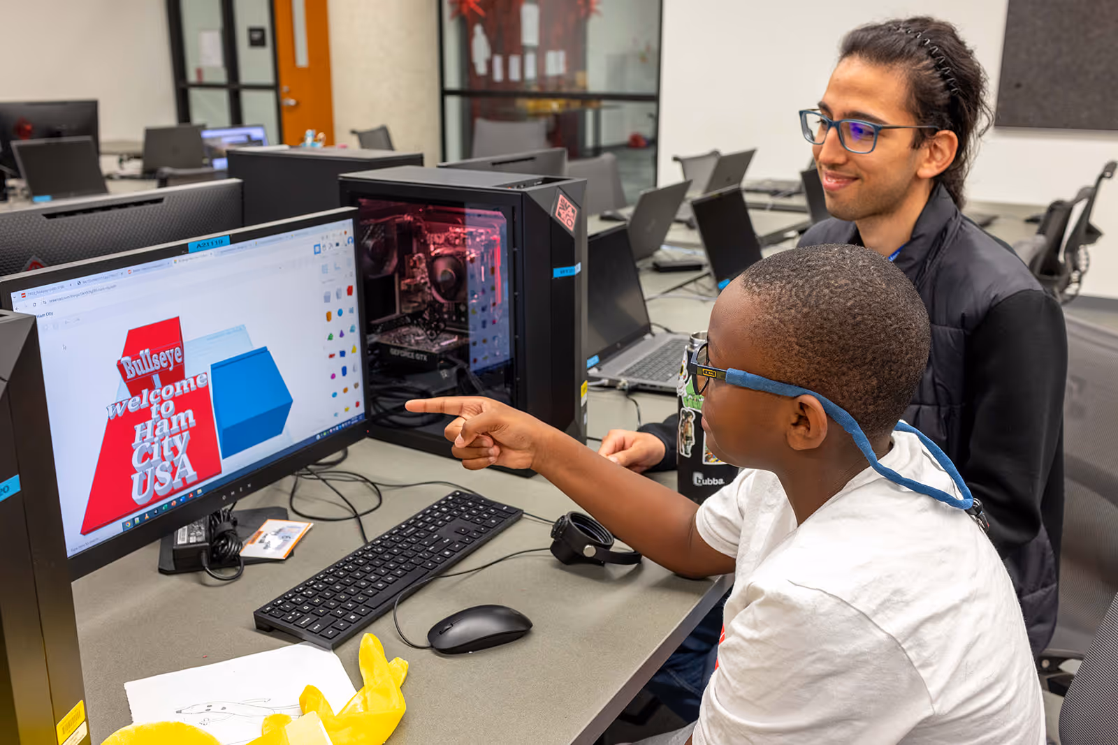 Two people at a computer workstation looking at a 3D modeling design on the monitor that says, 'Bullseye welcome to Ham City USA.'