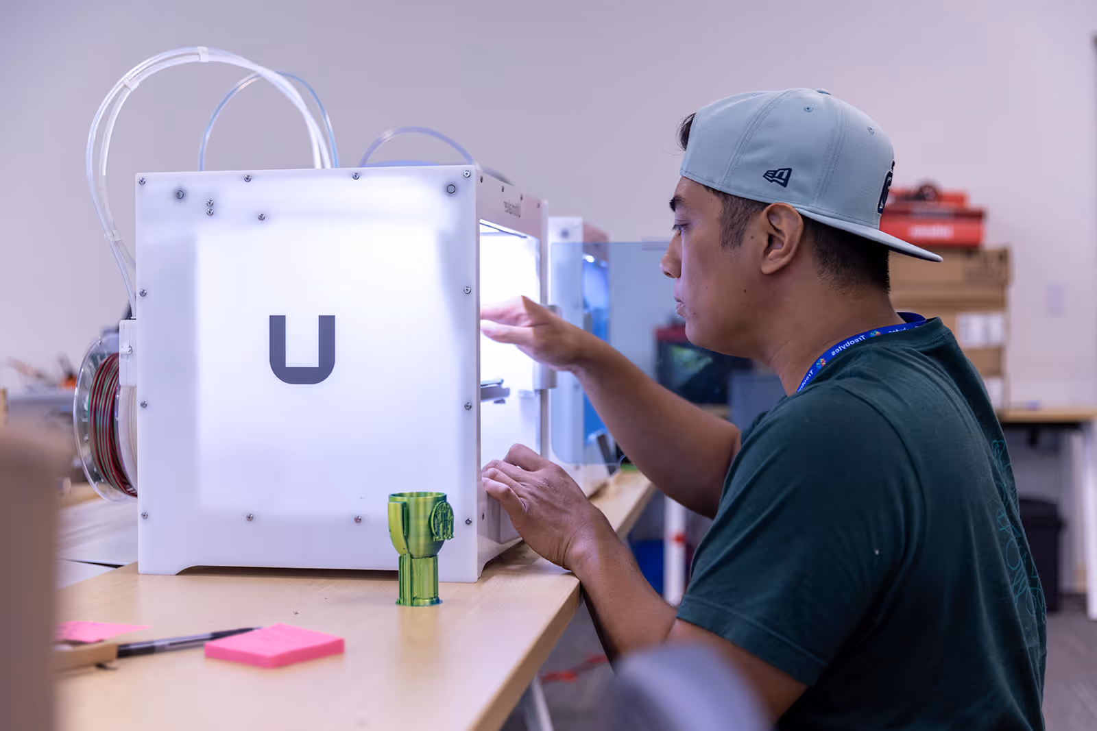 Person in a backward cap working on a 3D printer with a green 3D printed object on the table.