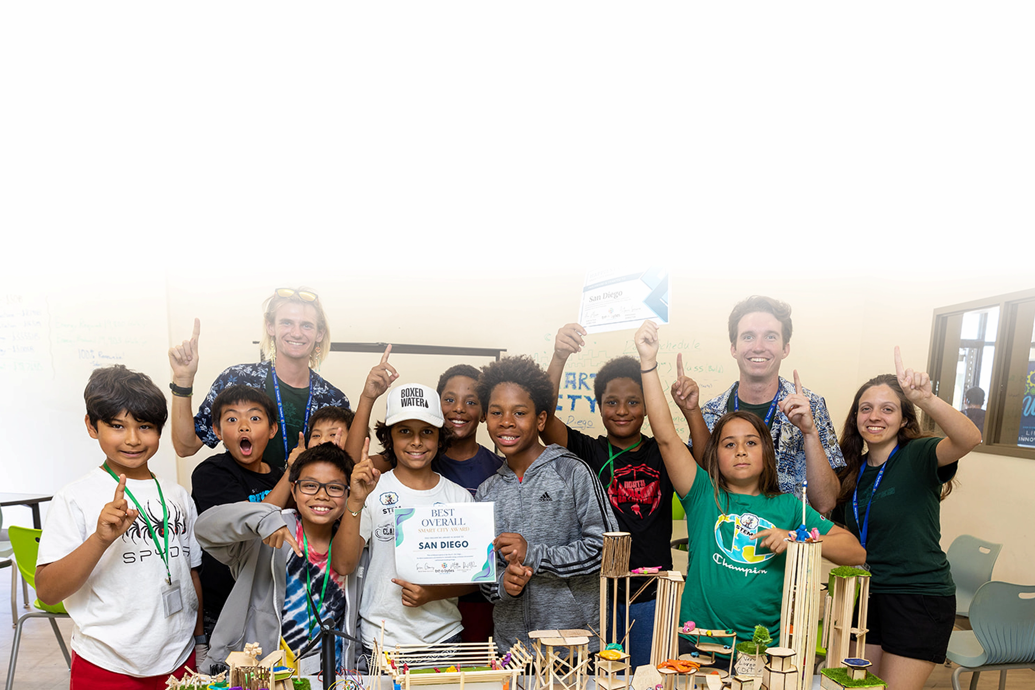A group of smiling children and two adults proudly hold certificates and pose behind a table with miniature architectural models.
