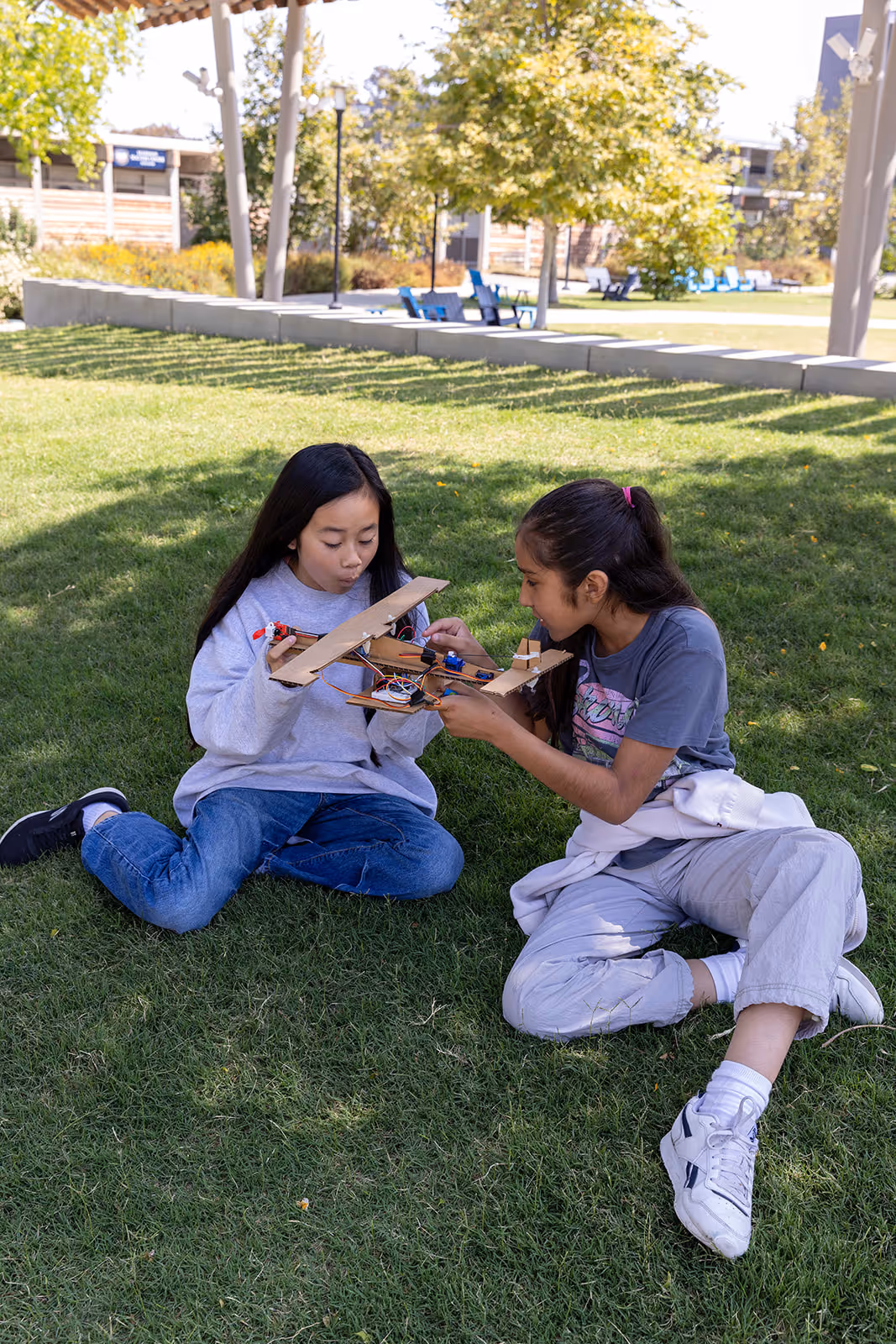 Two girls sitting on grass outdoors, focused on assembling or examining wooden model parts.