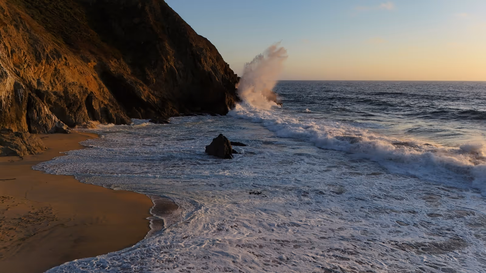 Ocean waves crashing against rocky cliffs at a sandy beach during sunset.